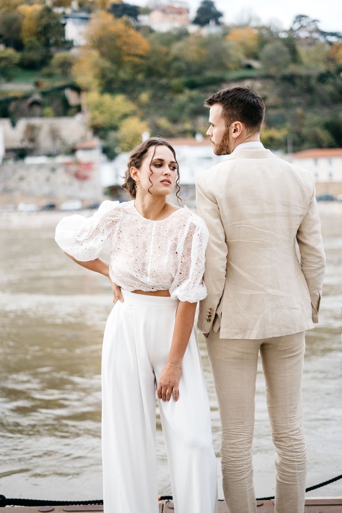 A couple stands by the water, the woman wearing a white lace blouse and high-waisted white pants, while the man is dressed in a light beige suit. They appear to be posing for a photo with a scenic background of trees and a distant building.